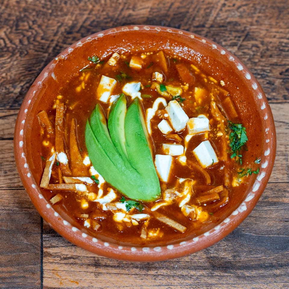 Overhead view of a terracotta bowl filled with red tortilla soup, garnished with avocado slices and cheese.