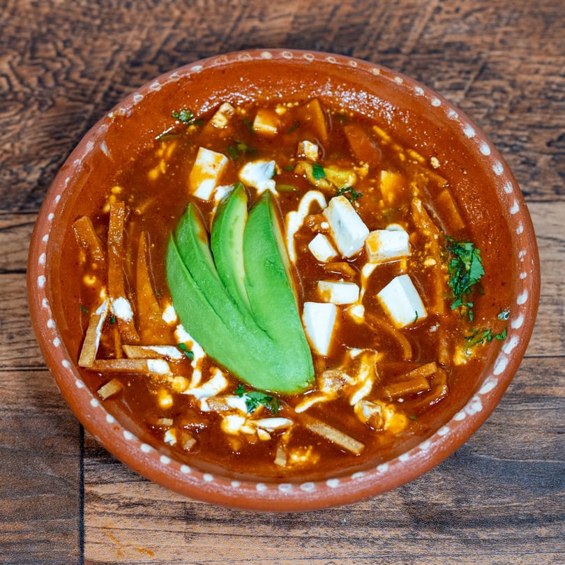 Overhead view of a terracotta bowl filled with red tortilla soup, garnished with avocado slices and cheese.