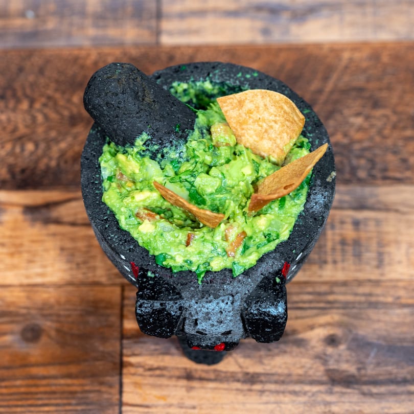 Fresh guacamole with tortilla chips and a pestle in a dark molcajete on a wooden table.