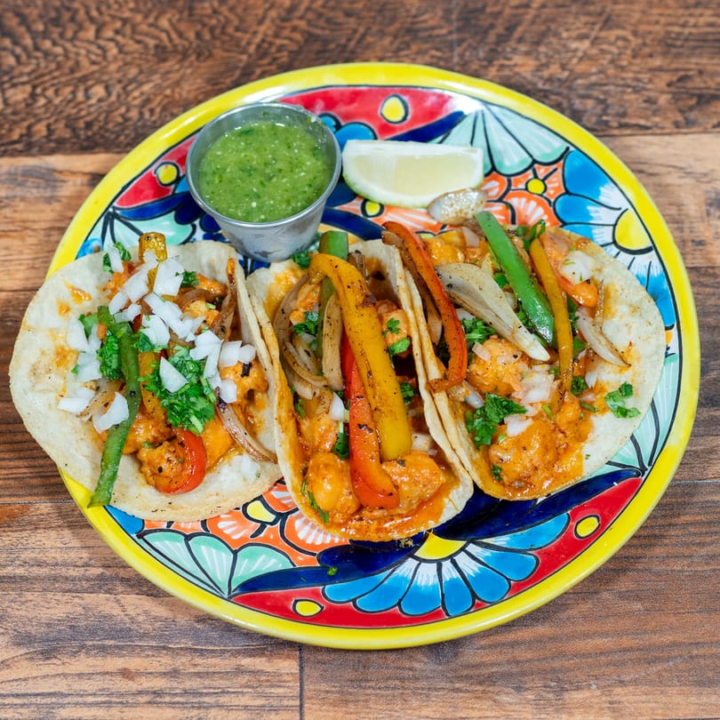 Three vibrant shrimp tacos with peppers, onions, and cilantro, served with salsa verde and lime.
