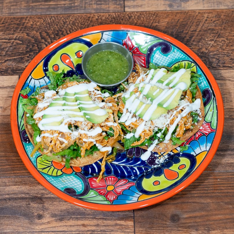 Two chicken tostadas with lettuce, avocado, crema, and salsa verde on a colorful plate.