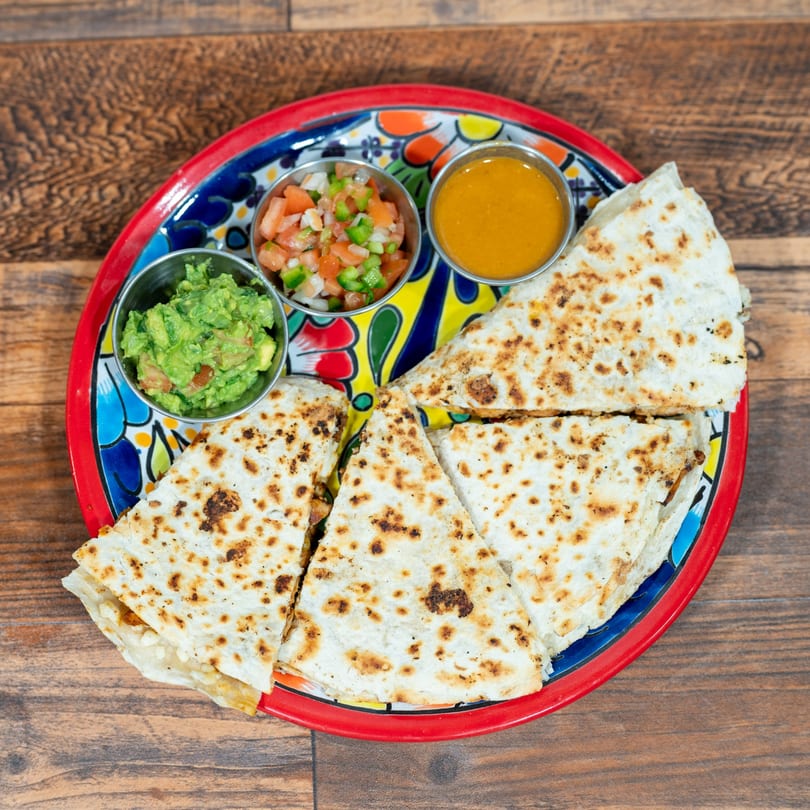 Quesadilla slices with sides of guacamole, pico de gallo, and orange sauce on a colorful plate.