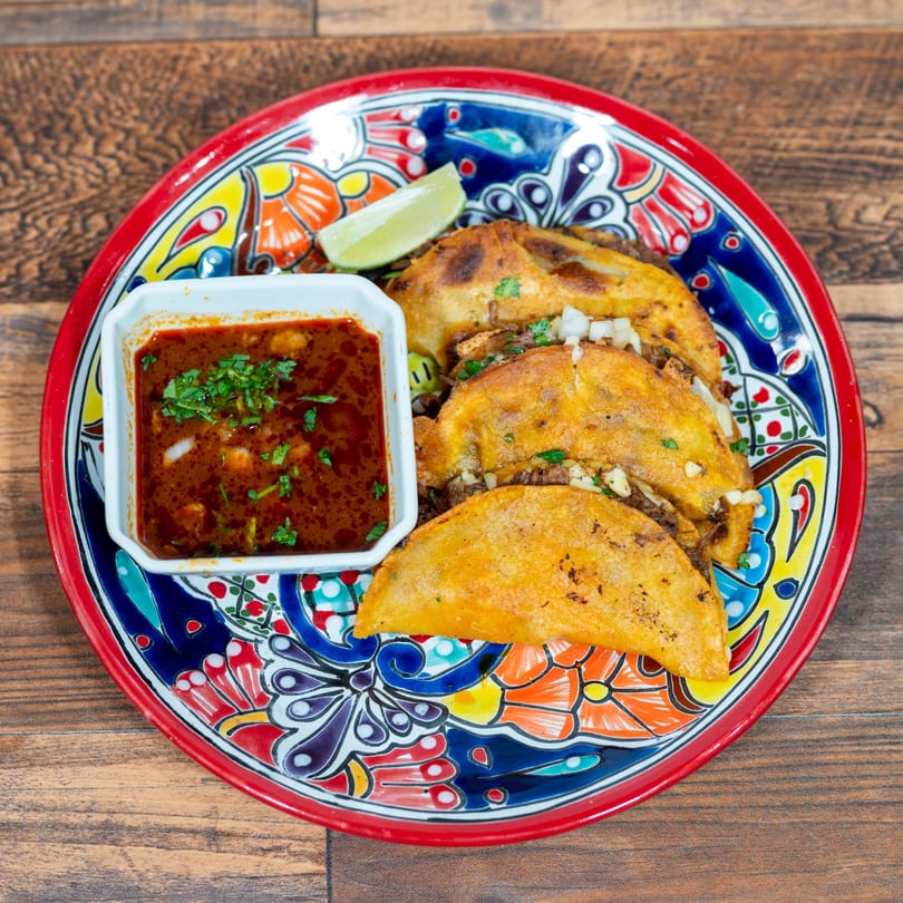 Four birria tacos with a side of consommé and a lime wedge on a colorful plate.