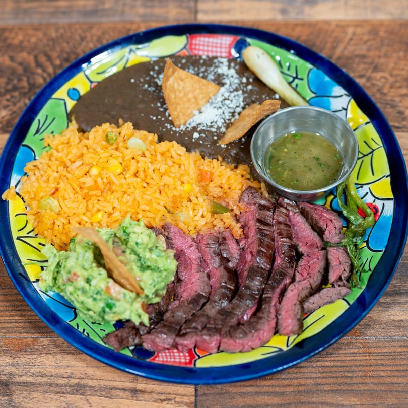 Sliced steak, rice, black beans, guacamole, and salsa on a colorful plate.