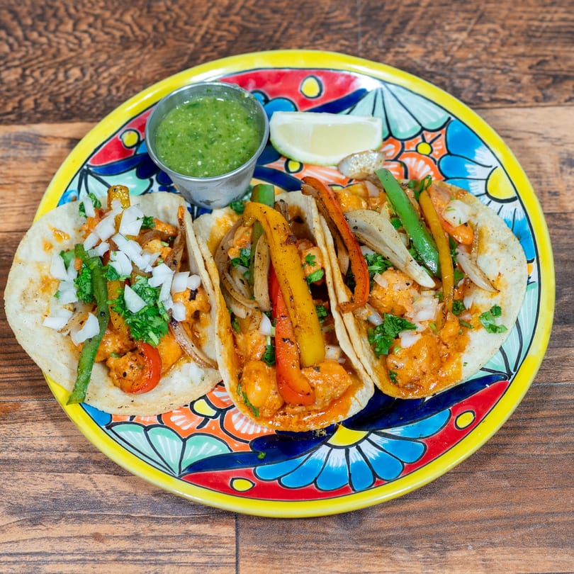 Three shrimp tacos with peppers, onions, cilantro, and salsa verde on a colorful plate.