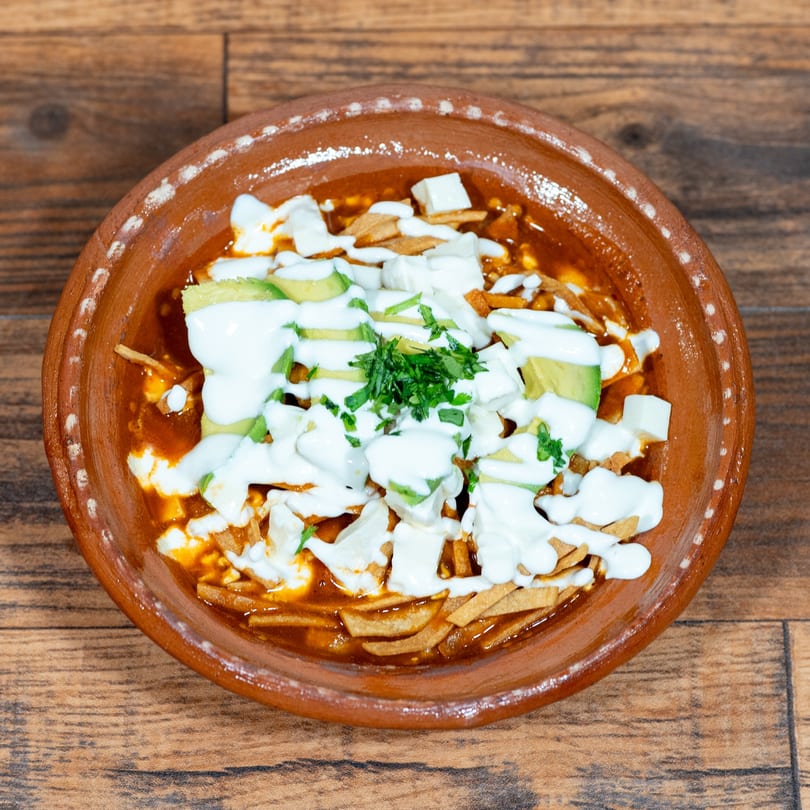 A close-up of a rustic clay bowl filled with a red stew, topped with avocado, cheese, and cream.