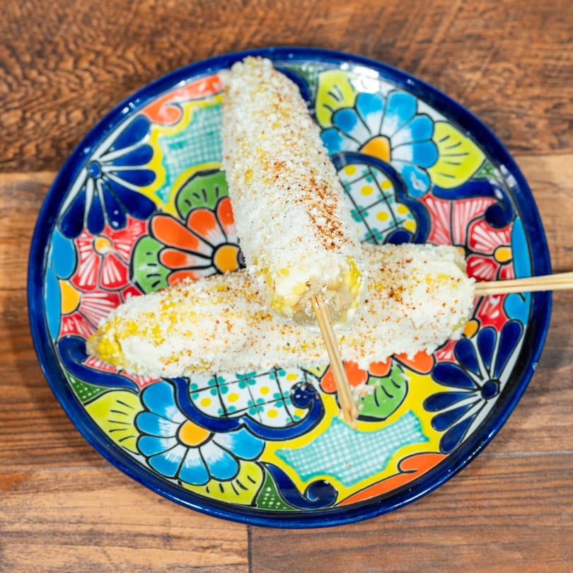 Two elotes, Mexican street corn, on a colorful floral plate on a wooden table.